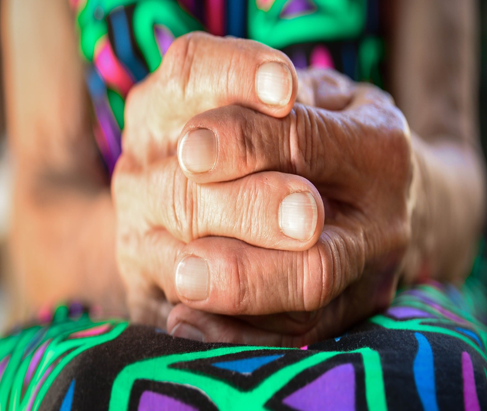 Closeup image of hands crossed together on a person's lap. The person is wearing an outfit with bright green, purple, blue, and pink colors. This Photo by Unknown Author is licensed under Creative Commons.