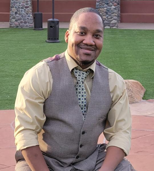 An African male who wearing a light brown shirt with a brown vest smiling for a picture