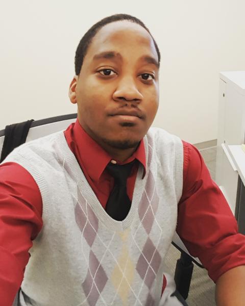 An young African American man with black hair, a mustache, a hint of a beard, a red polo shirt with a black tie, and a gray argyle sweater vest