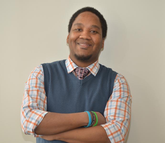 A picture of Kevin Wright who is an African American with black hair and goatee. Kevin is wearing an orange and light blue plaid shirt, a black and pink tie, and a dark navy blue vest.