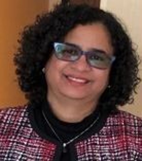 A Latina woman with black curly hair, glasses, a gold necklace, a black shirt, and one plaid cardigan