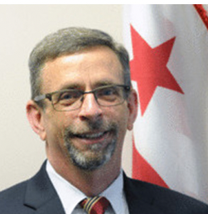 A white male adult with glasses, white hair and beard, blue suit with white collar shirt, and a plaid tie stands in front of the DC flag