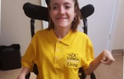 Amber Keohane, a young Caucasian woman with cerebral palsy, who has shoulder-length brown hair, black sunglasses, hoop earrings, and a yellow "DC Center for Living Independent" polo, sits in her wheelchair.